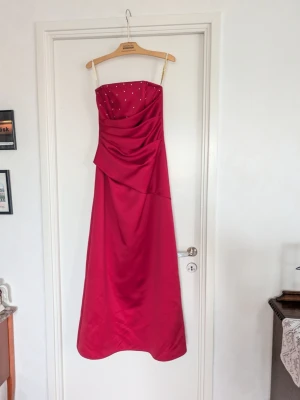 Röd balklänning - Elegant röd långklänning utan axelband och pärldetaljer upptill. Klänningen har draperad överdel och en figurnära passform upptill som faller ut i en rak kjol. Perfekt för bal eller festliga tillfällen.
