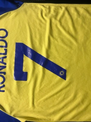 Gul och blå Al Nassr Ronaldo tröja - Säljer en gul och blå fotbollströja från Al Nassr med Ronaldo och nummer 7 på ryggen. Tröjan har klubbens logga och sponsor SHURFAH på framsidan, samt blå detaljer på axlarna. Perfekt för dig som vill representera laget och Ronaldo på planen.
