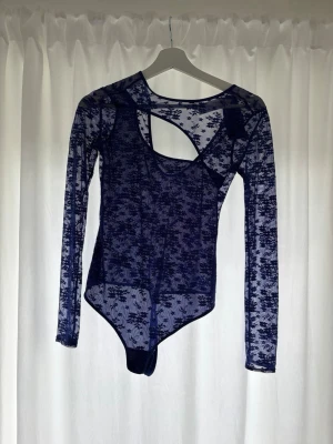 Blå spetsbody från hunkemöller  - Säljer en blå långärmad body från hunkemöller i transparent spets med blommönster och cool cut-out detalj vid axeln. Perfekt för dig som vill ha något unikt och trendigt. Materialet är mjukt och stretchigt, vilket gör den bekväm att bära. Prislappen är kvar och aldrig används