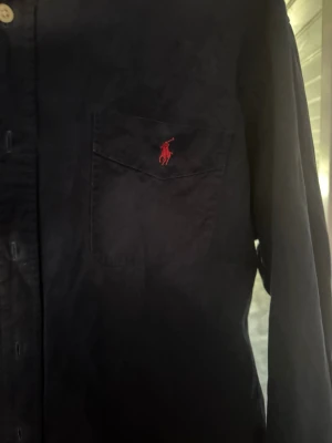 Mörkblå skjorta Ralph Lauren - Säljer en klassisk mörkblå skjorta från Ralph Lauren med röd broderad logga på bröstfickan. Skjortan har knappar framtill och långärmad design. Perfekt för dig som gillar stilrena plagg med ikonisk branding.