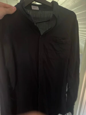 Svart skjorta från Houdini, XL - Klassisk svart skjorta från Houdini i storlek XL. Skjortan har lång ärm, bröstficka och knappar framtill. Perfekt för dig som gillar enkel och stilren design. Tillverkad i ett mjukt och bekvämt material.