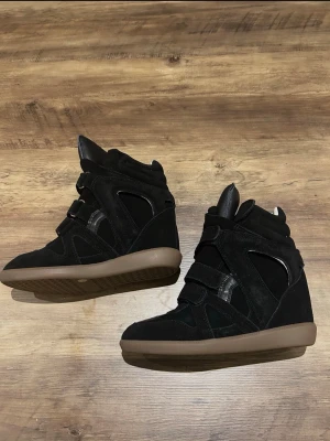 Svarta Isabel Marant skor - Snygga svarta wedge sneakers från Isabel Marant med högt skaft och kardborreband. Skorna har en mix av mocka och läderdetaljer samt en brun gummisula. Perfekta för dig som vill ha en sportig men ändå trendig look. Kommer ej med boxen 