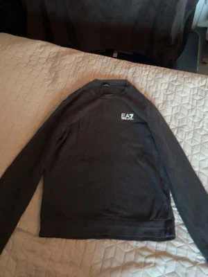 Svart sweatshirt från EA7 - Snygg svart sweatshirt från EA7 med rund halsringning och diskret logga på bröstet. Tröjan har långa ärmar och en klassisk passform, perfekt för en stilren och avslappnad look.