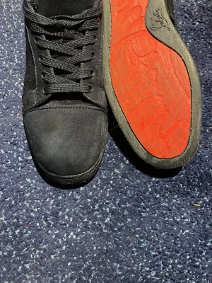 Svarta Christian Louboutin sneakers - Snygga svarta sneakers från Christian Louboutin med klassisk röd sula och diskret logga. Skorna har lågt skaft, svarta skosnören och är tillverkade i en mix av mocka och läder. Perfekta för dig som vill sticka ut med exklusiva detaljer och stilren design.