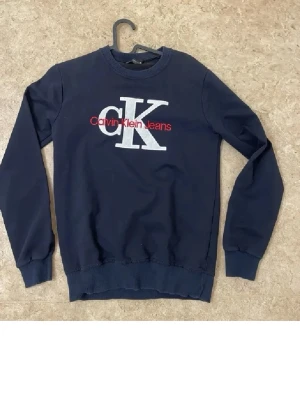 Mörkblå Calvin Klein sweatshirt - Mörkblå sweatshirt från Calvin Klein Jeans med stor vit CK-logga och röd text på bröstet. Tröjan har rund hals, långa ärmar och ribbade muddar vid ärmslut och nederkant. Perfekt för en avslappnad och stilren look.
