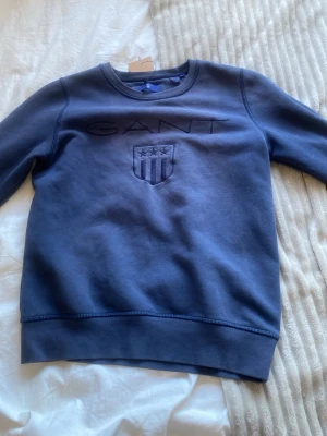 Mörkblå sweatshirt från GANT - Mörkblå sweatshirt från GANT med broderad logga och sköld på bröstet. Klassisk rund halsringning och ribbade muddar vid ärmslut och nederkant. Tillverkad i mjukt material för en bekväm känsla.