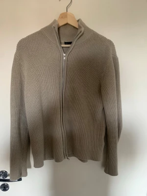 Beige ribbstickad ziptröja 157 - Beige ribbstickad tröja från 157 med dragkedja framtill och hög krage. Tröjan har lång ärm och är tillverkad i ett mjukt, grovstickat material som ger en avslappnad och stilren look.