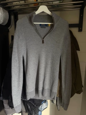 Grå stickad tröja Polo Ralph Lauren - Snygg grå stickad tröja från Polo Ralph Lauren i tvättbar merinoull. Tröjan har halv dragkedja vid halsen, ribbade muddar och en klassisk passform. Perfekt för lager-på-lager och stilrena outfits.