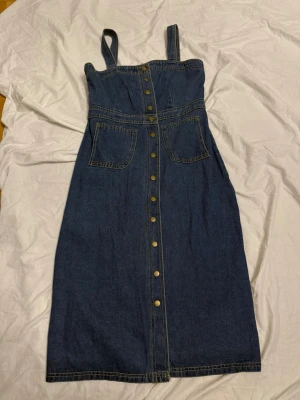 Jeansklänning med knappar och fickor - Snygg jeansklänning i blå denim med breda axelband, två stora fickor framtill och knappar längs hela framsidan. Klänningen har markerad midja och kontrastsömmar som ger en cool look. Perfekt för dig som gillar en avslappnad men trendig stil.