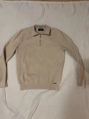 Beige stickad tröja med halvzip ICON - Snygg beige stickad tröja från ICON med halvzip och krage. Tröjan har ribbad struktur, långa ärmar och en liten metallbricka vid nederkanten. Perfekt för dig som gillar stilrena och enkla plagg med en modern touch. Tröjan är lite sliten vid axeln som man ser på den tredje bilden, dock syns det inte om man inte rör vid det.