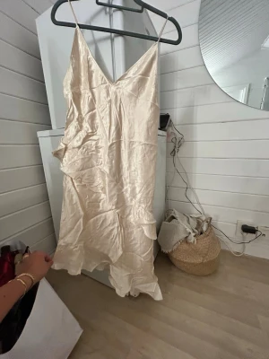 Champagnefärgad satin klänning M - Champagnefärgad kort klänning i satin med smala axelband och v-ringning. Klänningen har volangdetaljer och en lätt glansig finish som ger en lyxig känsla. Perfekt för dig som gillar en elegant och trendig stil. Är inte använd o har fortfarande kvar prislapp