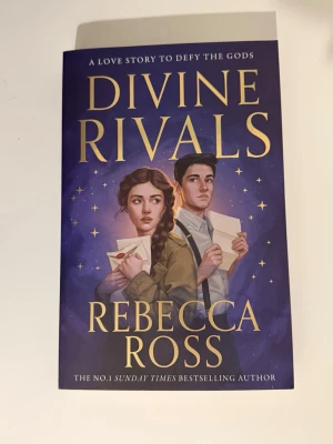 Divine Rivals av Rebecca Ross - En magisk och romantisk ungdomsroman med lila omslag och illustration av två unga personer som håller brev. Boken handlar om Iris och Roman som knyts samman genom brev och magi i en värld där gudar och krig hotar deras öde. Helt ny skick, aldrig läst.