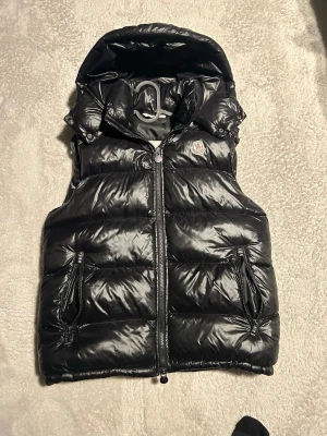 Svart dunväst från Moncler - Snygg svart dunväst från Moncler med glansig finish, två dragkedjefickor och hög krage med huva. Klassisk Moncler-logga på bröstet och quiltad design. Perfekt för lager på lager och streetwear-vibe.