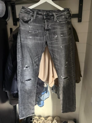 Jack & Jones Grå slitna jeans med raka ben  - Säljer ett par grå jeans med slitningar och hål på flera ställen. Jeansen har raka ben, klassisk femficksmodell och knappgylf. Perfekta för en avslappnad och trendig streetstil.