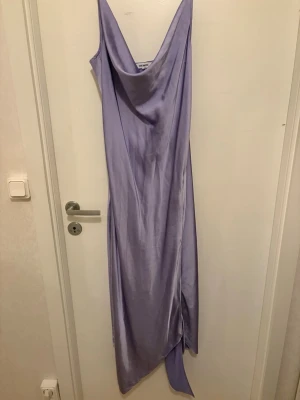 Pastel lila satinklänning från Steve Madden - Pastel lila lång satinklänning från Steve Madden. Klänningen har en draperad urringning och asymmetrisk nederkant som ger en cool och trendig look. Perfekt för dig som gillar stilrena och eleganta plagg med lite extra edge.