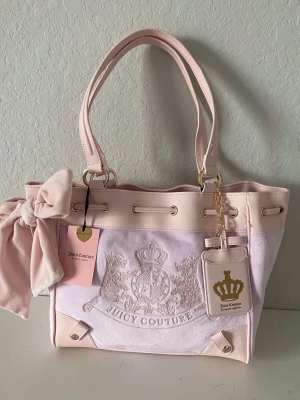 Ljusrosa daydreamer scottie tote Juicy Couture - Superfin ljusrosa handväska från Juicy Couture med broderad logga framtill, stora handtag och dekorativ rosett på sidan. Väskan har guldfärgade detaljer, en tag med kronmotiv och är gjord i mjukt tyg och konstläder.