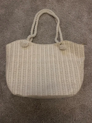 Beige strandväska  - Snygg strandväska i beige stickat material med dekorativa rep som handtag. Väskan har ett rymligt foder i ljus textil och stängs med dragkedja. Perfekt för dig som gillar en avslappnad och trendig stil.