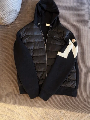 Svart Moncler hoodie med vadderad front - Mycket bra skick! Säljer en svart pufferjacka från Moncler med stickade ärmar och huva. Jackan har ett quiltat mönster på framsidan, vit detalj på ena ärmen och Moncler-logga på axeln. Säljer då jag inte använder den längre.