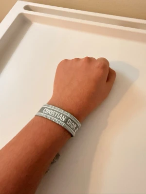 Grått armband / dekorativt snöre – Dior-inspirerat, nytt - Stilrent grått armband/snöre med dekorativa tofsar och vävd text. Kan bäras som armband eller användas som accessoar/dekoration (t.ex. på väska).  🔹 Nytt & oanvänt 🔹 Levereras i plastpåse 🔹 Justerbar storlek