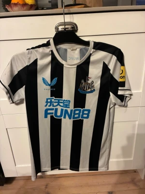 Newcastle United matchtröja Isak 14 - Snygg Newcastle United matchtröja med svarta och vita ränder, sponsortryck i blått och klubbmärke på bröstet. På ryggen står det ISAK och nummer 14 i rött. Korta ärmar och rund hals. Perfekt för fotbollsfans! Använd ett par gånger och har storleken S