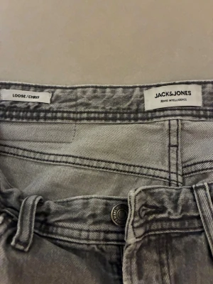 Grå loose fit jeans från Jack & Jones - Säljer ett par grå loose fit jeans från Jack & Jones, modell Chris. Jeansen har klassisk femficksdesign, bälteshällor och knappgylf. Perfekta för en avslappnad och trendig look. Ett  hål på höger knä.