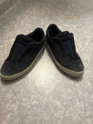 Svarta sneakers i mocka från Axel Arigato - Snygga svarta sneakers från Axel Arigato i mjuk mocka med vit sula. Klassisk låg modell utan snörning och diskret logga på sidan. Perfekta för en clean och stilren look.