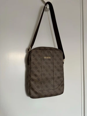 Brun IPad väska från Guess - Snygg IPad väska från Guess i brunt konstläder med logomönster över hela väskan och guldfärgad logga framtill. Väskan har en justerbar axelrem i mörkbrunt och dragkedja upptill.