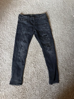 Svarta Slitna Jack & Jones jeans  - Säljer ett par svarta skinny jeans med slitningar och hål på framsidan. Jeansen har klassisk femficksdesign, låg midja och smal passform. Perfekta för en edgy och trendig stil. Materialet är denim med stretch för extra komfort. Pris kan diskuteras vid snabb affär!