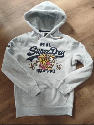 Grå Superdry hoodie med tigertryck - Snygg ljusgrå hoodie från Superdry med stort tryck framtill som visar en tiger, blommor och japanska tecken. Hoodien har huva med dragsko, känguruficka och ribbade muddar. Perfekt för en avslappnad streetstyle-look.