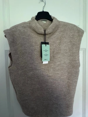 Beige stickad väst från Gina Tricot - Mysig beige stickad väst med hög krage från Gina Tricot. Tillverkad i en mjuk ull- och alpackablandning som ger en lyxig känsla. Perfekt att styla över en skjorta eller t-shirt för en trendig lager-på-lager-look.