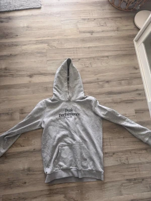 Grå hoodie från Peak Performance - Snygg grå hoodie från Peak Performance med svart logga broderad på bröstet. Klassisk känguruficka framtill och stor huva. Tillverkad i mjukt sweatshirtmaterial som är perfekt för chill dagar. Enkel och stilren design som funkar till det mesta.