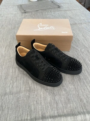 Svarta Louboutin sneakers med nitar - Snygga svarta sneakers från Christian Louboutin med mattsvarta nitar på tån och sidorna. Skorna har en mix av mocka och läder, svarta skosnören och en klassisk låg siluett. Perfekta för dig som vill sticka ut med en edgy vibe.