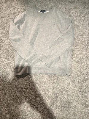Grå sweatshirt från Polo Ralph Lauren - Klassisk grå sweatshirt från Polo Ralph Lauren med rund halsringning och liten mörkblå logga broderad på bröstet. Tröjan har ribbade muddar vid ärmslut och nederkant. Perfekt basplagg i mjukt material. Storlek XL 18-20 på barn/ungdom sortimentet vilket ungefär motsvarar en lite bredare S
