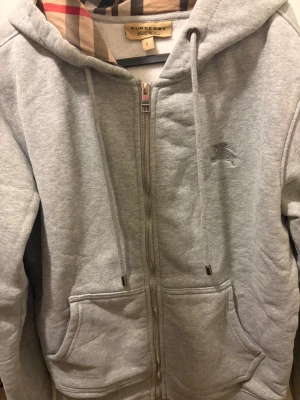 Grå zip-hoodie från Burberry - Snygg grå hoodie från Burberry med dragkedja, klassiskt rutigt foder i huvan och broderad logga på bröstet. Mjuk bomullsblandning, två stora fickor fram och justerbar huva med snören. Perfekt för en chill och stilren look. Sitter litet rekommenderat på folk som är 170-175
