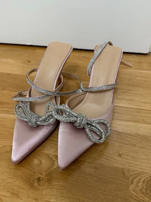 Rosa pumps med strass och rosett - Snygga spetsiga pumps i ljusrosa satin med höga klackar. Skorna har tunna remmar dekorerade med strass och en stor glittrig rosett framtill. Perfekta för att sticka ut och ge extra bling till din outfit.