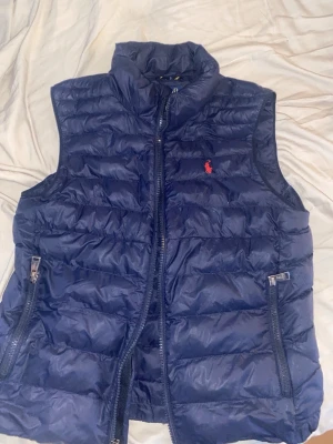 Mörkblå dunväst från Ralph Lauren - Snygg mörkblå dunväst från Ralph Lauren med hög krage och dragkedja framtill. Västen har två fickor med dragkedja och den klassiska röda Polo-loggan broderad på bröstet. Perfekt för lager-på-lager och riktigt skön att bära.