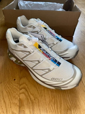 Salomon XT-6 - Salomon XT-6 sneakers i vitt med grå detaljer och färgglada logoband på plösen. Skorna har en sportig design med mesh och syntetmaterial, snabbsnörning och robust sula. Perfekta för dig som gillar streetwear och outdoor-vibe.