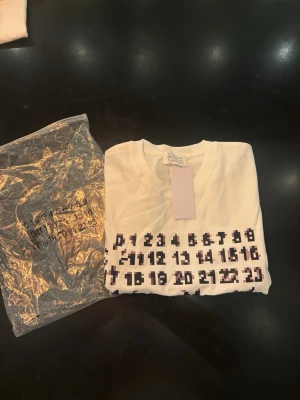 Maison Margiela t-shirt  - Vit t-shirt från Maison Margiela med svart och pixlat tryck framtill som visar siffror och logotyp. Klassisk rund halsringning och korta ärmar. Perfekt för dig som gillar unika designer och streetwear-vibe.