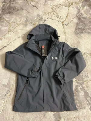 Mörkgrå vindjacka från Under Armour - Snygg mörkgrå vindjacka från Under Armour med huva och dragkedja. Jackan har två fickor med dragkedja, elastiska muddar och reflekterande detaljer på bröstet. Perfekt för blåsiga dagar och sportiga outfits.