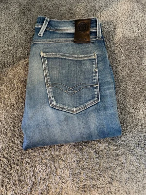 Blåa jeans med slitningar från Replay - Säljer ett par blå jeans från Replay med slitningar och trasiga detaljer på framsidan. Jeansen har klassisk femficksdesign, ljus tvätt och dekorativa sömmar på bakfickan. Perfekta för en avslappnad och trendig look. Modellen är anbass😁