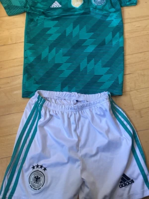 Tysklands matchställ Adidas - Säljer ett Tysklands matchställ från Adidas med grön tröja i grafiskt mönster och vita shorts med gröna ränder. Både tröjan och shortsen har Tysklands landslagslogga och Adidas-logga. Perfekt för fotbollsträning eller match.