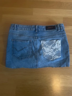 Blå jeanskjol med spetsdetalj ONLY - Kort blå jeanskjol från ONLY med rå kant nertill och femficksmodell. Ena bakfickan har vit spetsdetalj med blommönster som ger kjolen en unik touch. Klassisk knapp och dragkedja framtill.