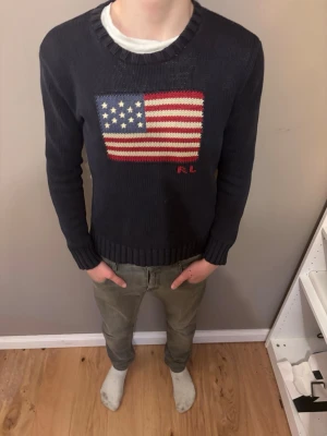 Ralph lauren tröja - Mörkblå stickad tröja från Ralph Lauren med amerikansk flagga i rött, vitt och blått på bröstet samt RL-broderi. Tröjan har rund halsringning och ribbade muddar vid ärmslut och nederkant. Perfekt för dig som gillar klassisk preppy stil.