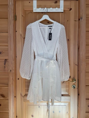 Vit omlottklänning från Boohoo - Söt vit kortklänning från Boohoo med omlottmodell, volangdetaljer och knytband i midjan. Klänningen har långa, transparenta ärmar med prickigt mönster och v-ringad hals. Perfekt för studenten eller sommarens alla tillfällen☀️🎉