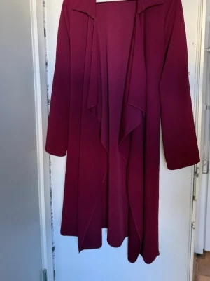 Vinröd lång kappa med vattenfallsfront - Säljer en vinröd kappa med snygg vattenfallsfront och långa ärmar. Kappan har en enkel, stilren design och en liten silverfärgad knappdetalj på ärmen. Materialet känns mjukt och följsamt, perfekt för dig som gillar en clean look.