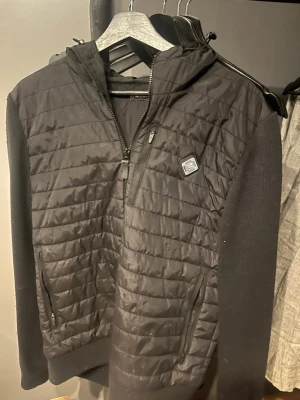 Svart vindjacka från J.Lindeberg - Snygg svart Cardigan från J.Lindeberg med quiltad front, stickade ärmar och huva. Jackan är i storlek L men passar M. Den har en liten spricka baktill som är enkelt reparationsbar,men är i övrigt i väldigt bra skick.