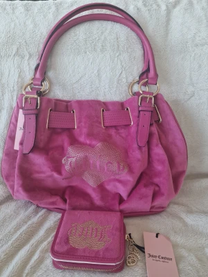 Rosa Juicy Couture handväska med plånbok - Snygg rosa handväska från Juicy Couture i mjuk mockaliknande material med glittrande Juicy-logga i strass framtill. Väskan har dubbla handtag med guldfärgade detaljer och matchande plånbok med samma logga och hjärtdetalj. Perfekt för dig som gillar statement-accessoarer.