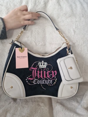 Juicy Couture svart och vit handväska - Juicy Couture handväska i svart sammet med vita detaljer i läderimitation. Väskan har guldfärgade kedjor, broderad logga med strass och krona samt en matchande liten vit accessoarväska framtill. Perfekt för dig som gillar statement-väskor och trendig design.