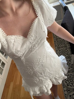 Vit kort klänning med broderade blommor - Supersöt vit kort klänning med puffärmar, volangkant nertill och broderade blommor över hela tyget. Klänningen har en fyrkantig ringning med dekorativa kanter och är perfekt för sommarens alla tillfällen. Passar perfekt till student, helt ny med prislappen kvar, storlek 38💕