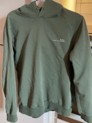 Grön hoodie från A.P.C. - Snygg grön hoodie från A.P.C. med minimalistisk design och liten vit logga på bröstet. Hoodien har huva, långa ärmar och ribbade muddar. Perfekt för en avslappnad och stilren look. Passar S